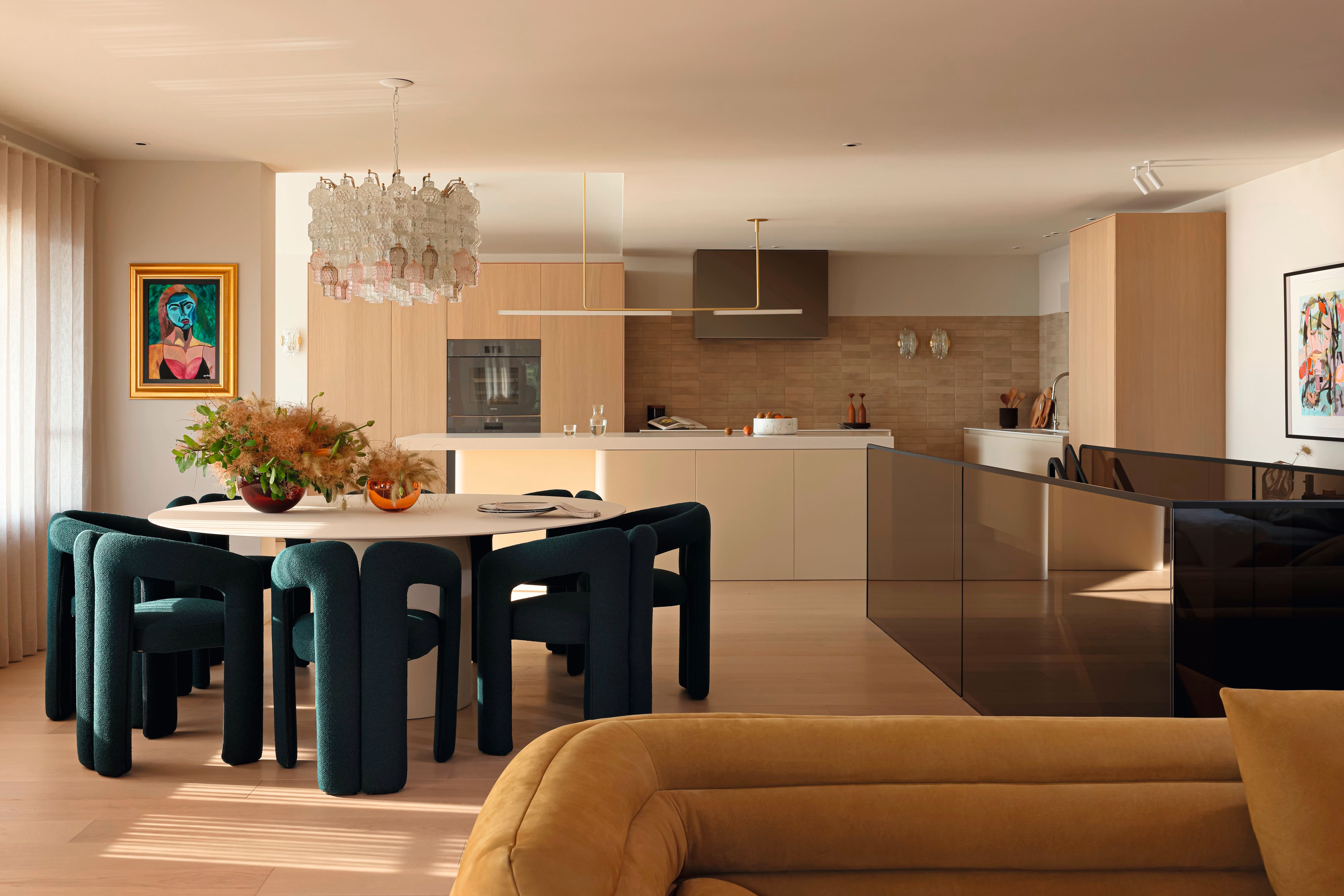 A modern open‑plan kitchen and dining area with green upholstered chairs, a round table, and light wood cabinetry.