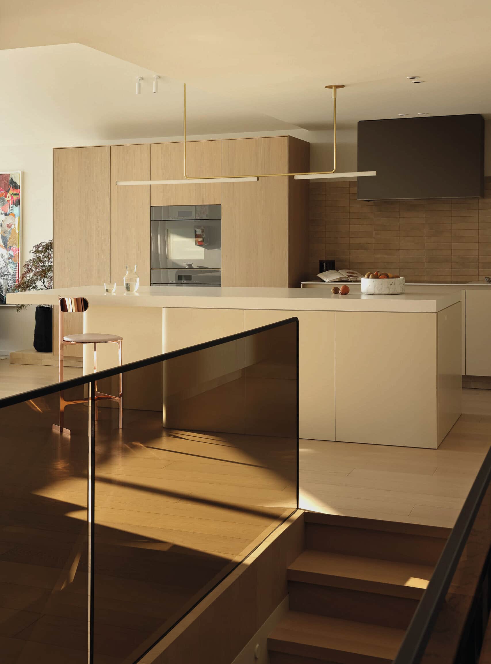 A modern kitchen with light wood cabinetry, a large island, and warm sunlight reflecting off a glass stair railing.