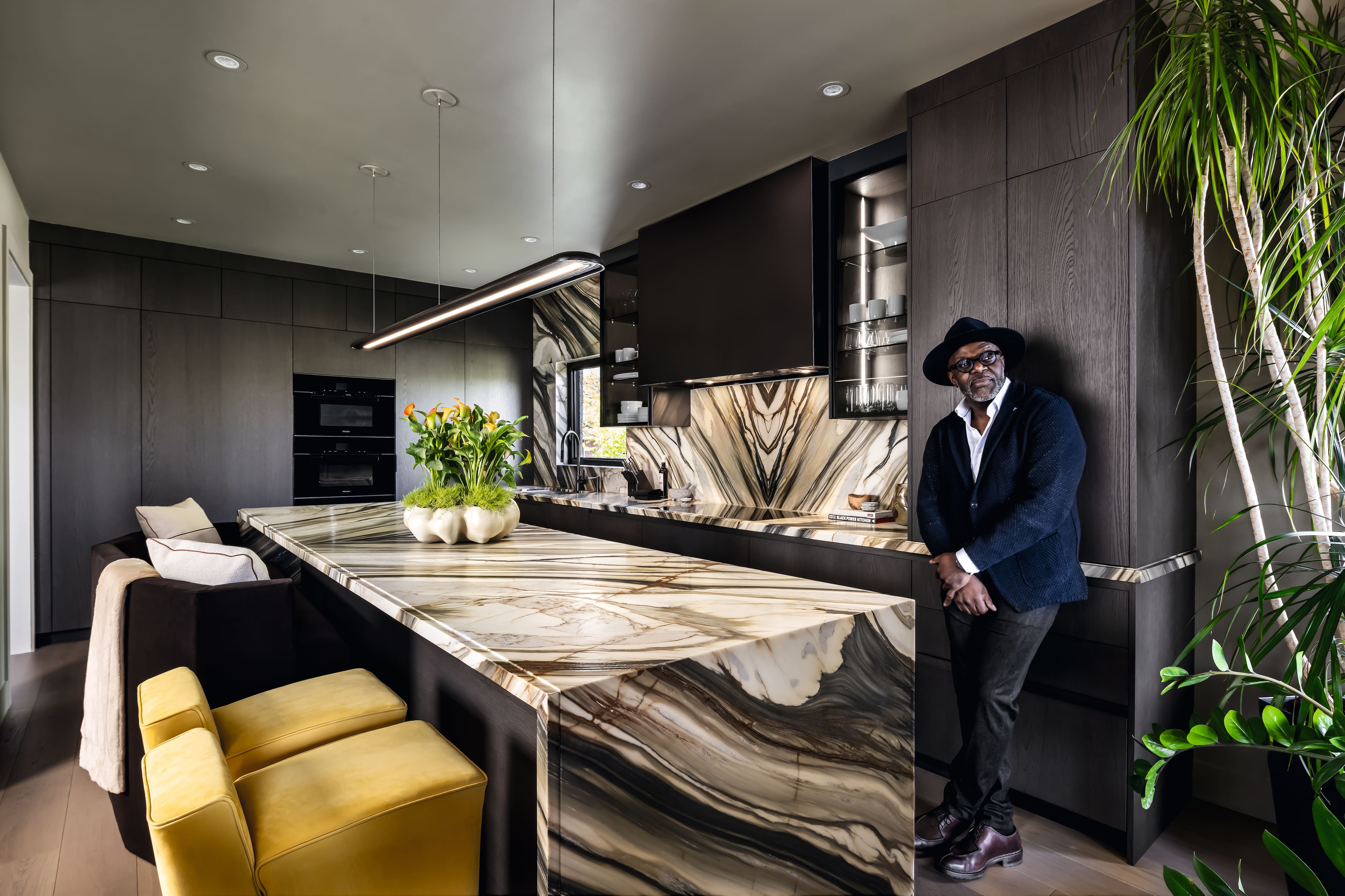 A modern kitchen with dark cabinetry and a long island featuring dramatic stone countertops. Upholstered seating lines one side of the island, while a person stands casually at the far end, leaning against the counter in a suit jacket.