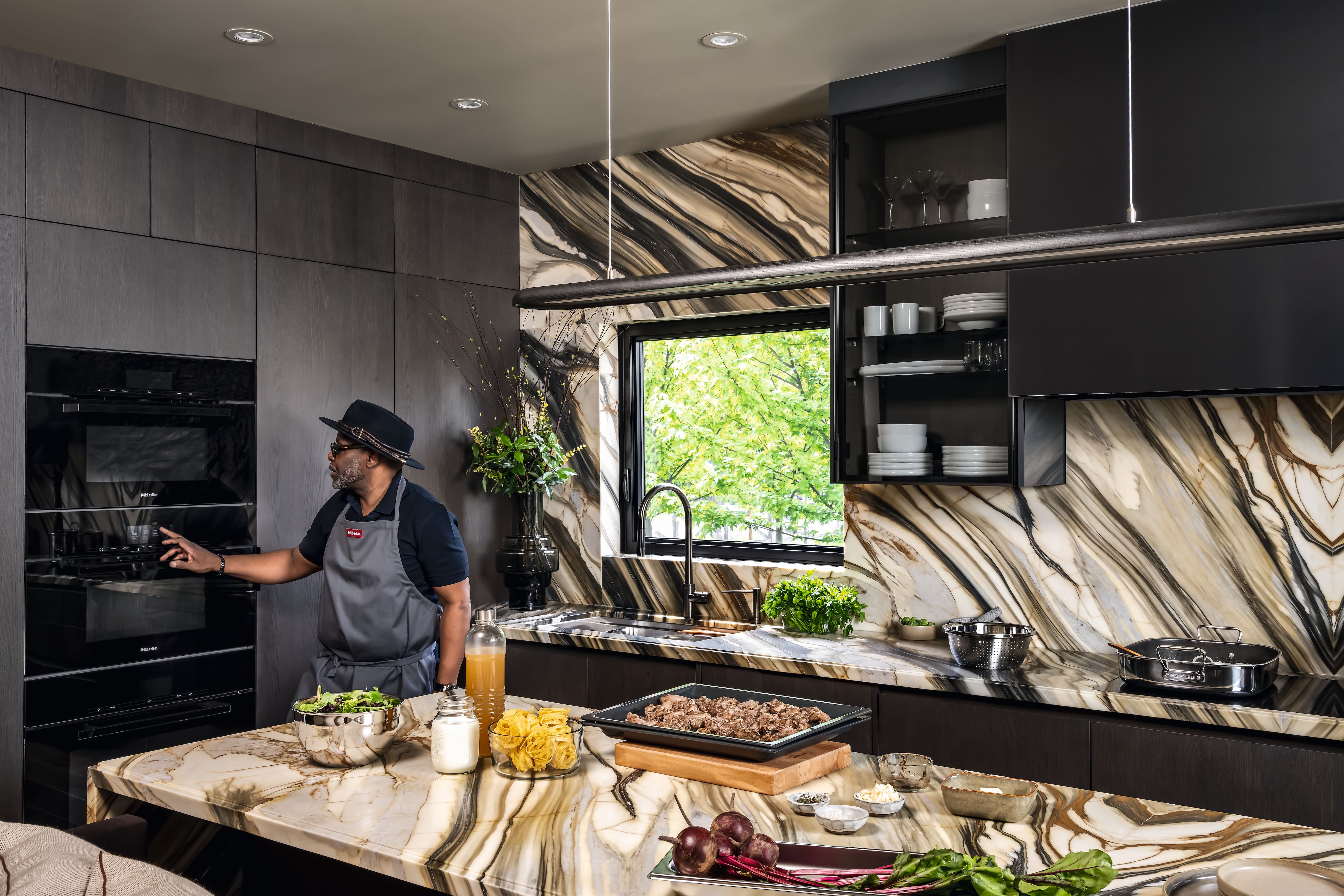 A person prepares food at a marble-patterned kitchen island with dark cabinetry and a built-in oven.