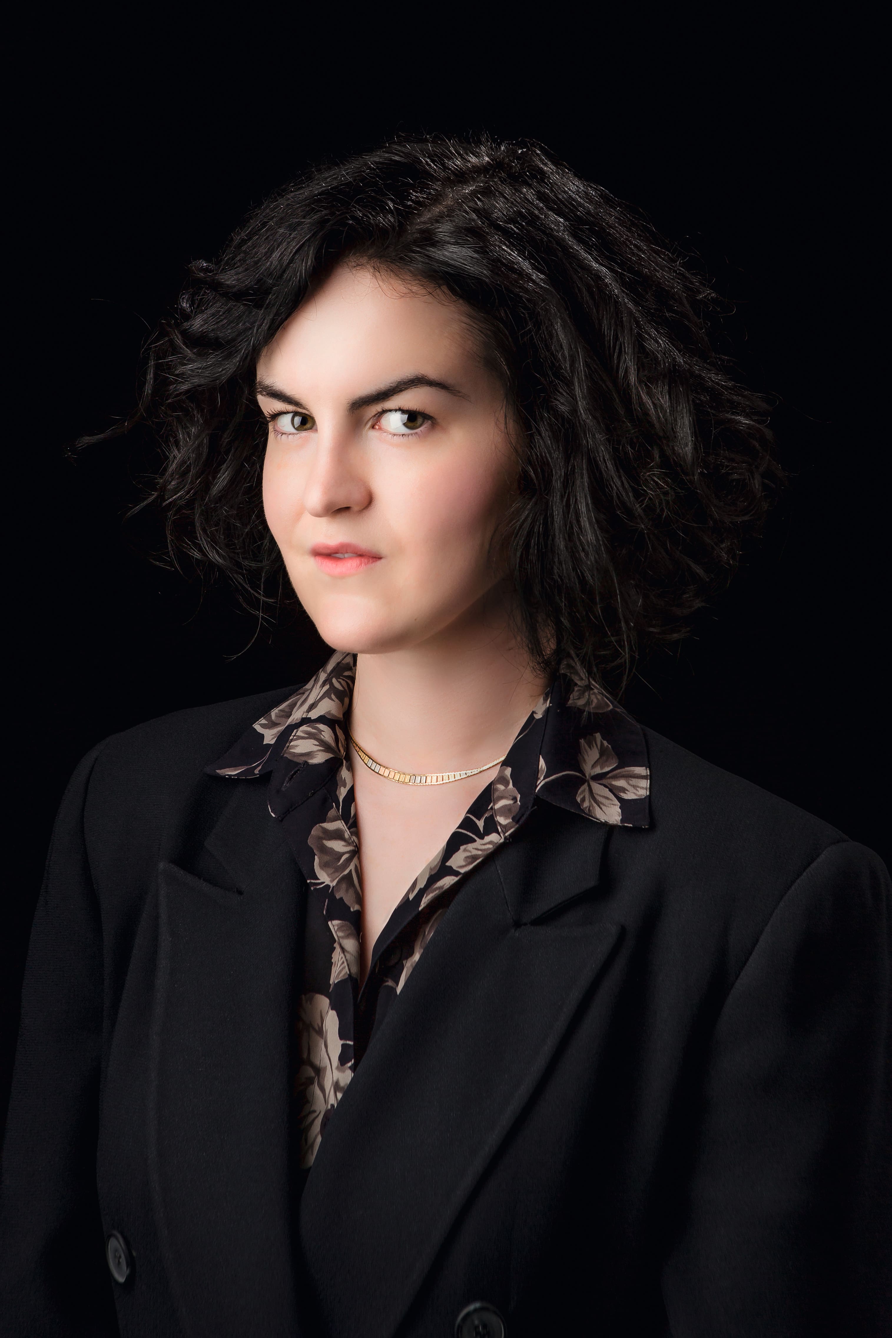 A portrait of a person with short, wavy dark hair, wearing a dark blazer over a patterned shirt and a delicate necklace, set against a solid black background.