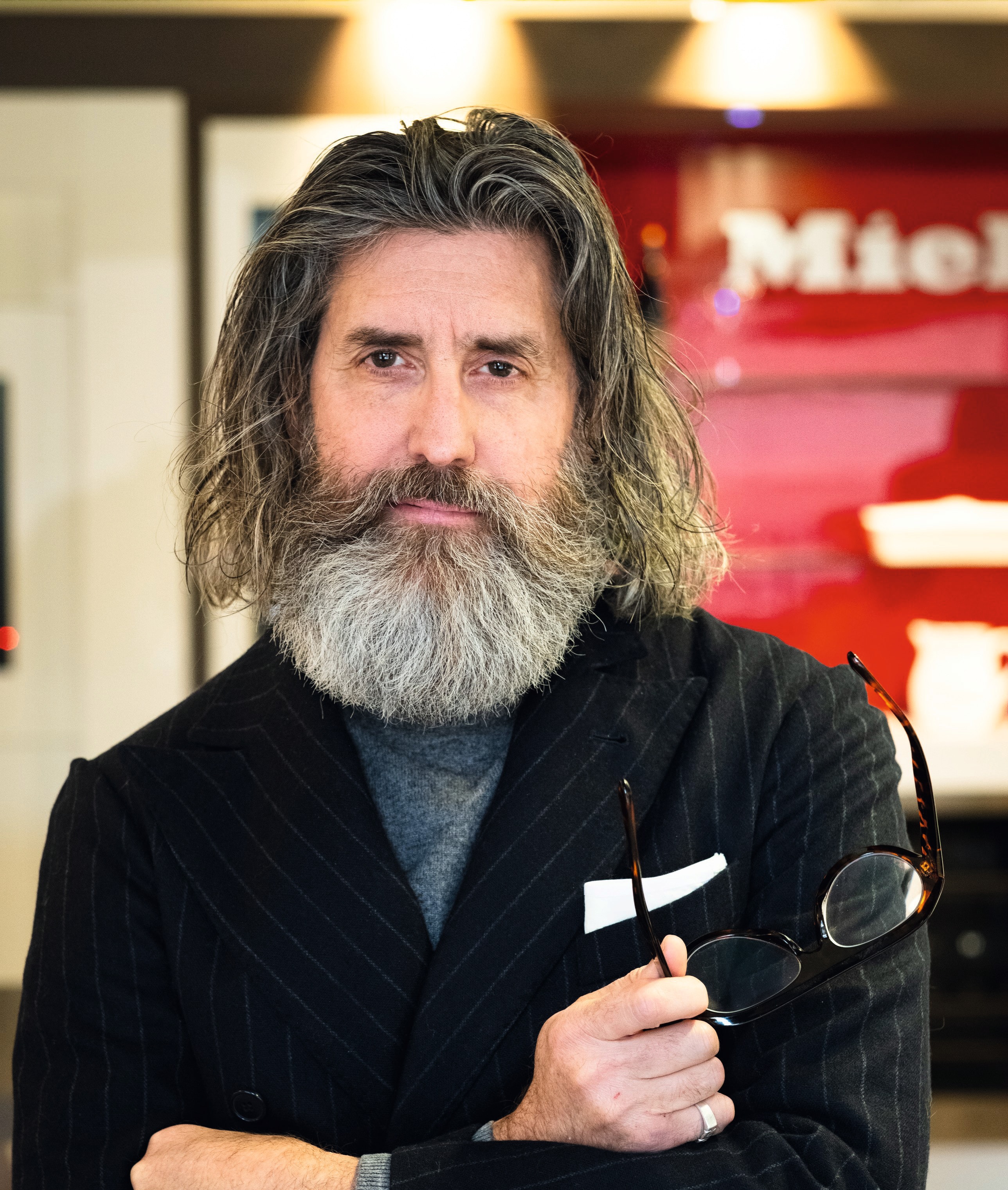 A portrait of a person with long, wavy gray hair and a full gray beard, wearing a dark pinstripe blazer over a high‑neck shirt and holding a pair of eyeglasses, standing indoors with a red and white background.