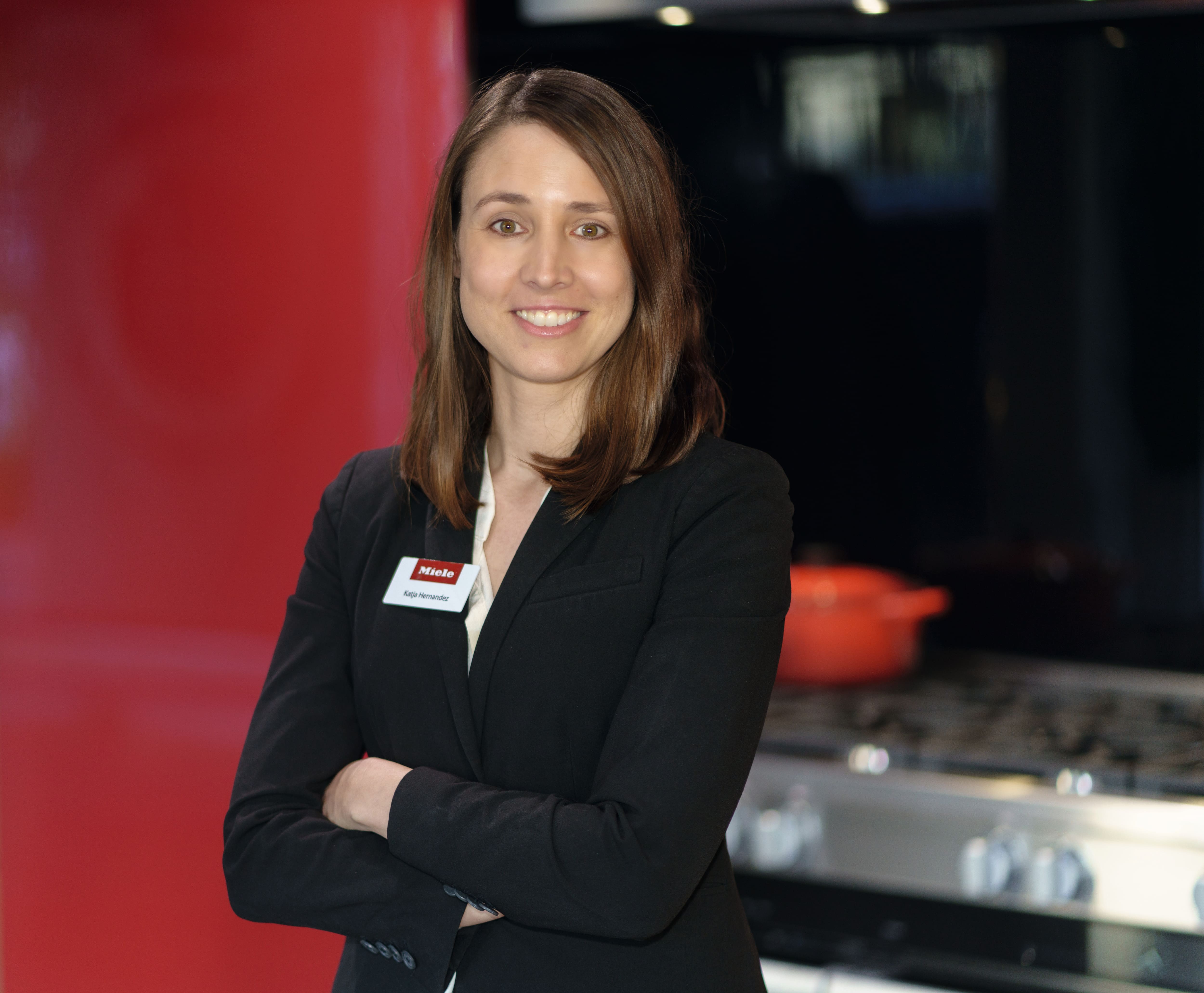 A portrait of a person with shoulder‑length brown hair wearing a black blazer and white shirt, standing with arms crossed in a modern kitchen showroom with a red wall and stainless‑steel appliances in the background.