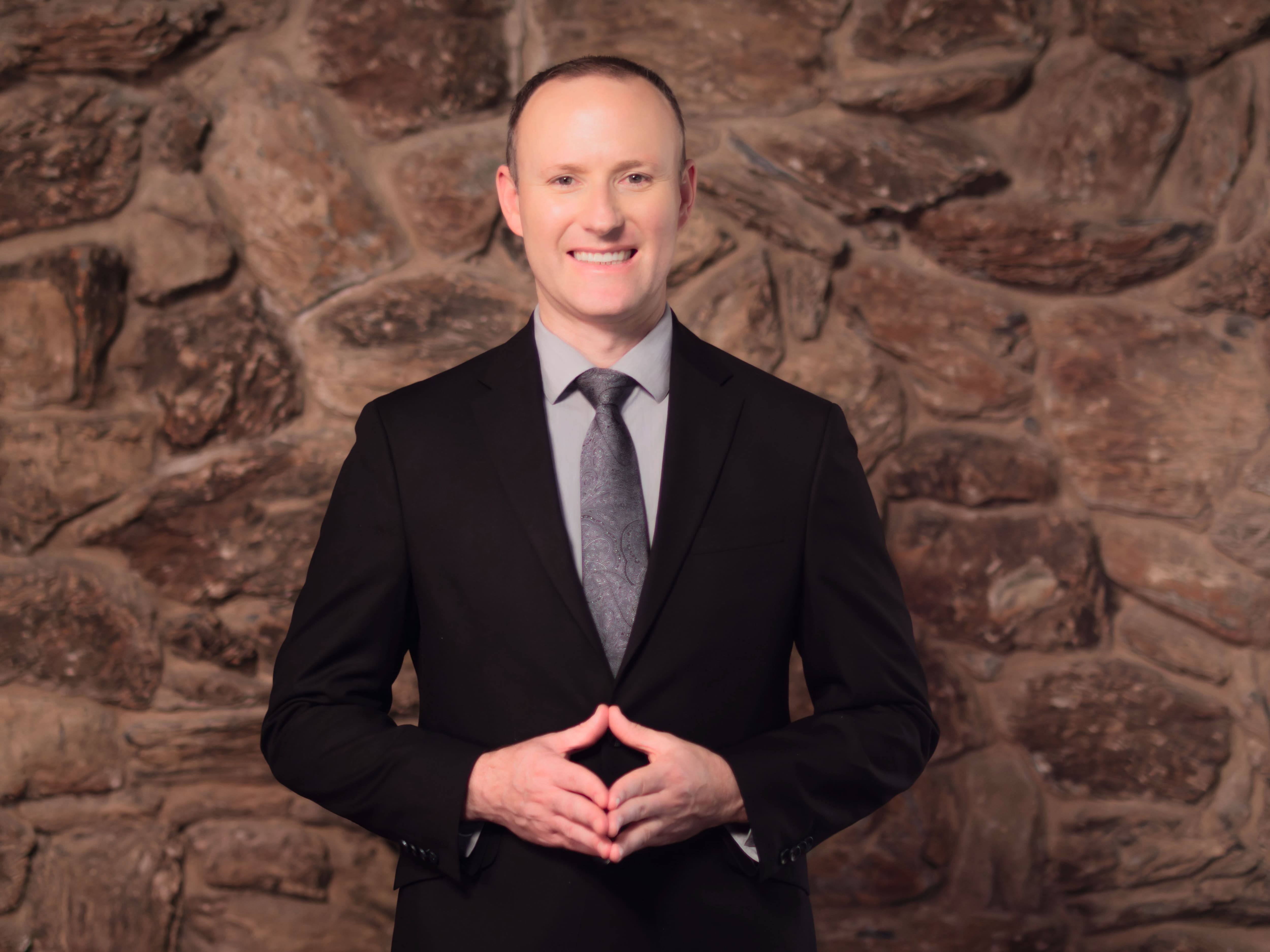 A portrait of a person wearing a dark suit, light dress shirt, and patterned tie, standing against a textured stone wall with hands loosely clasped at the waist.