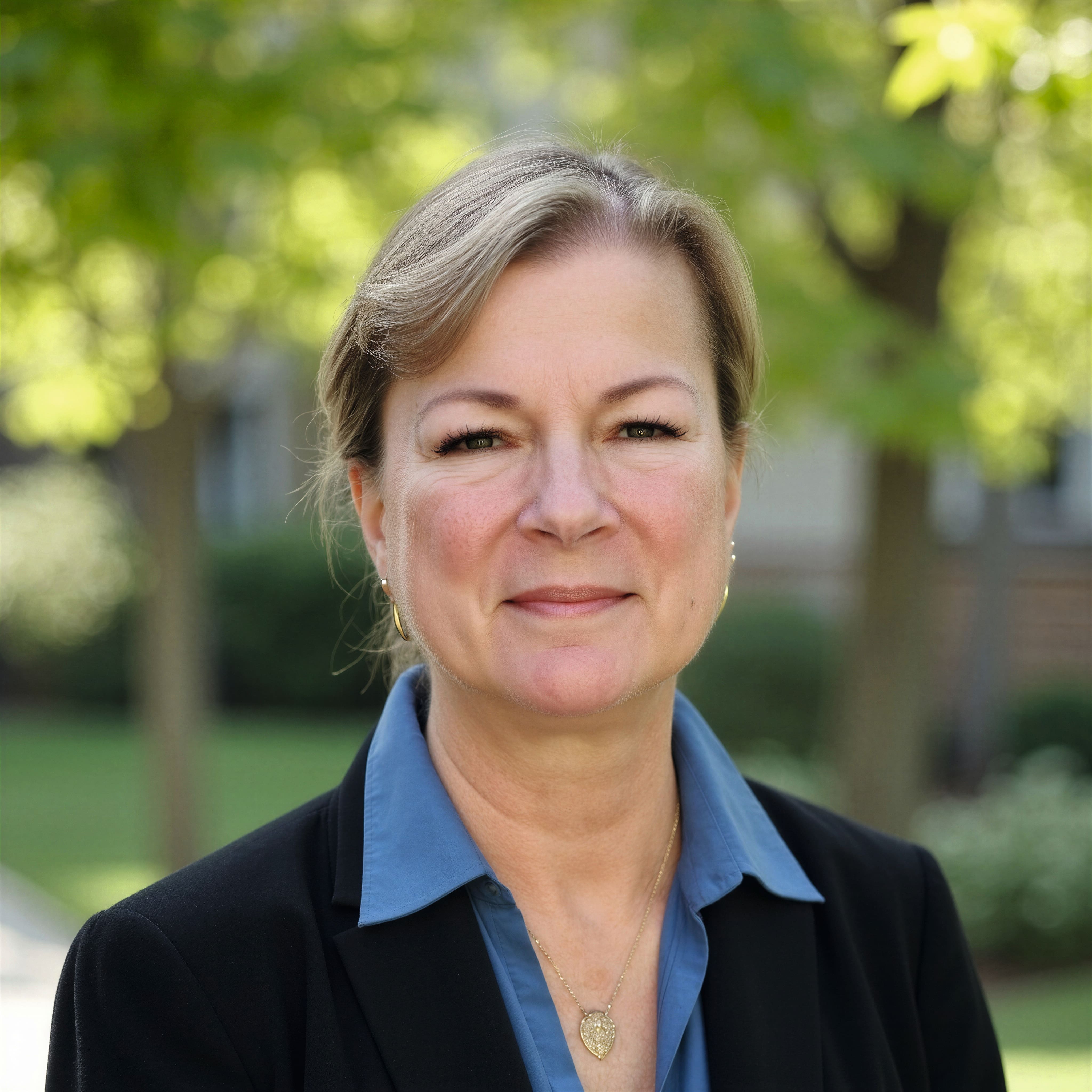 A portrait of a person with light brown hair pulled back, wearing a blue collared shirt under a dark blazer and a gold pendant necklace, standing outdoors in a green, tree‑filled area.