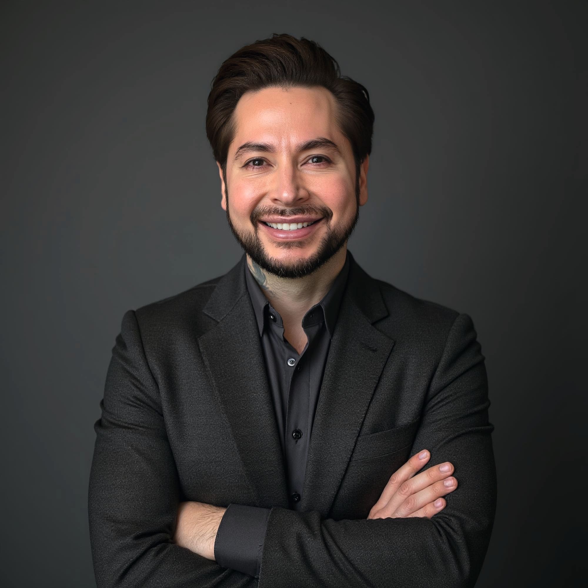 A portrait of a person with neatly styled dark brown hair wearing a dark suit jacket over a button‑down shirt, standing against a plain gray background with arms crossed.