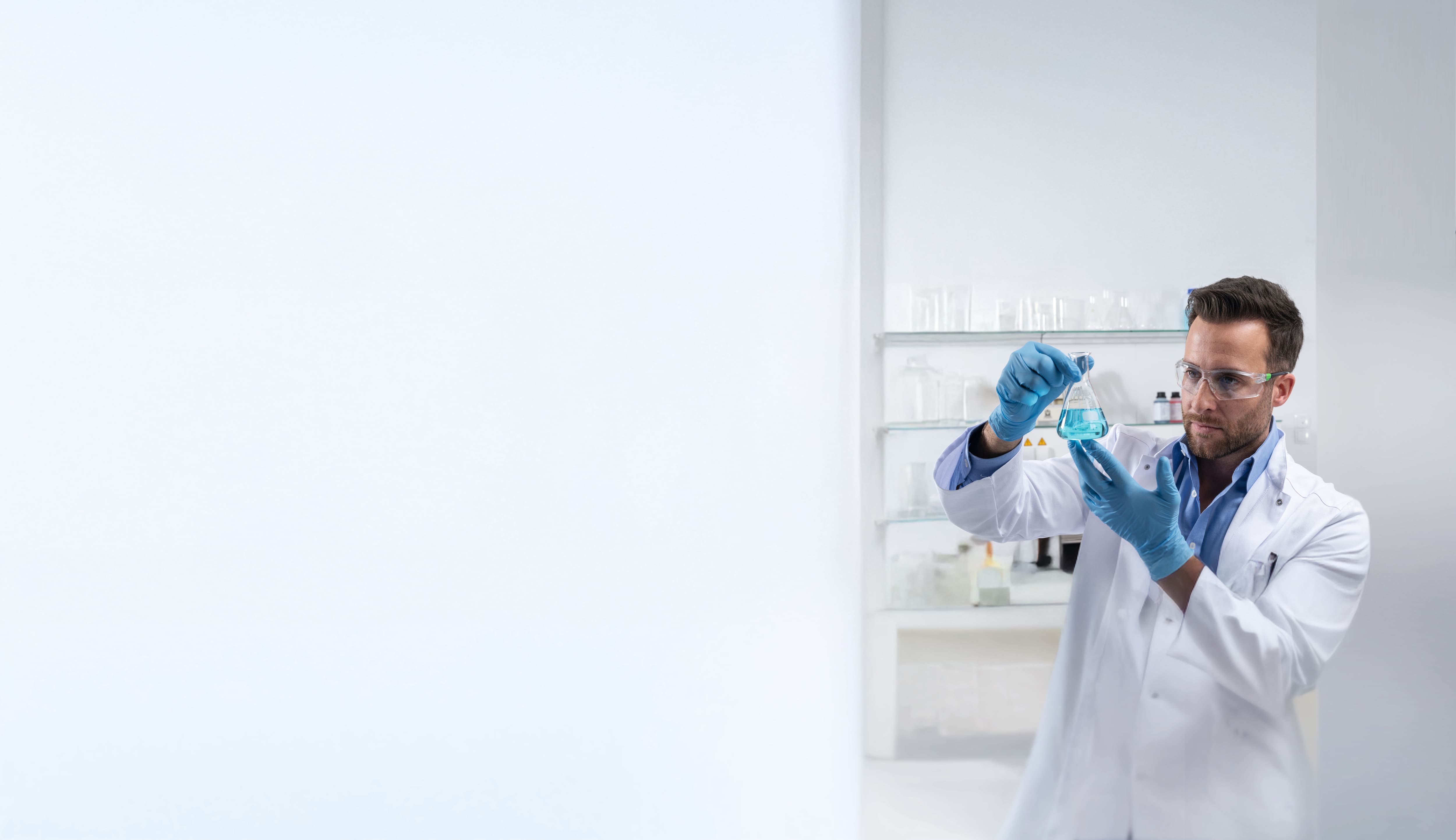 A laboratory assistant dressed in a white lab coat, gloves and safety glasses is holding an Erlenmeyer flask containing blue liquid.