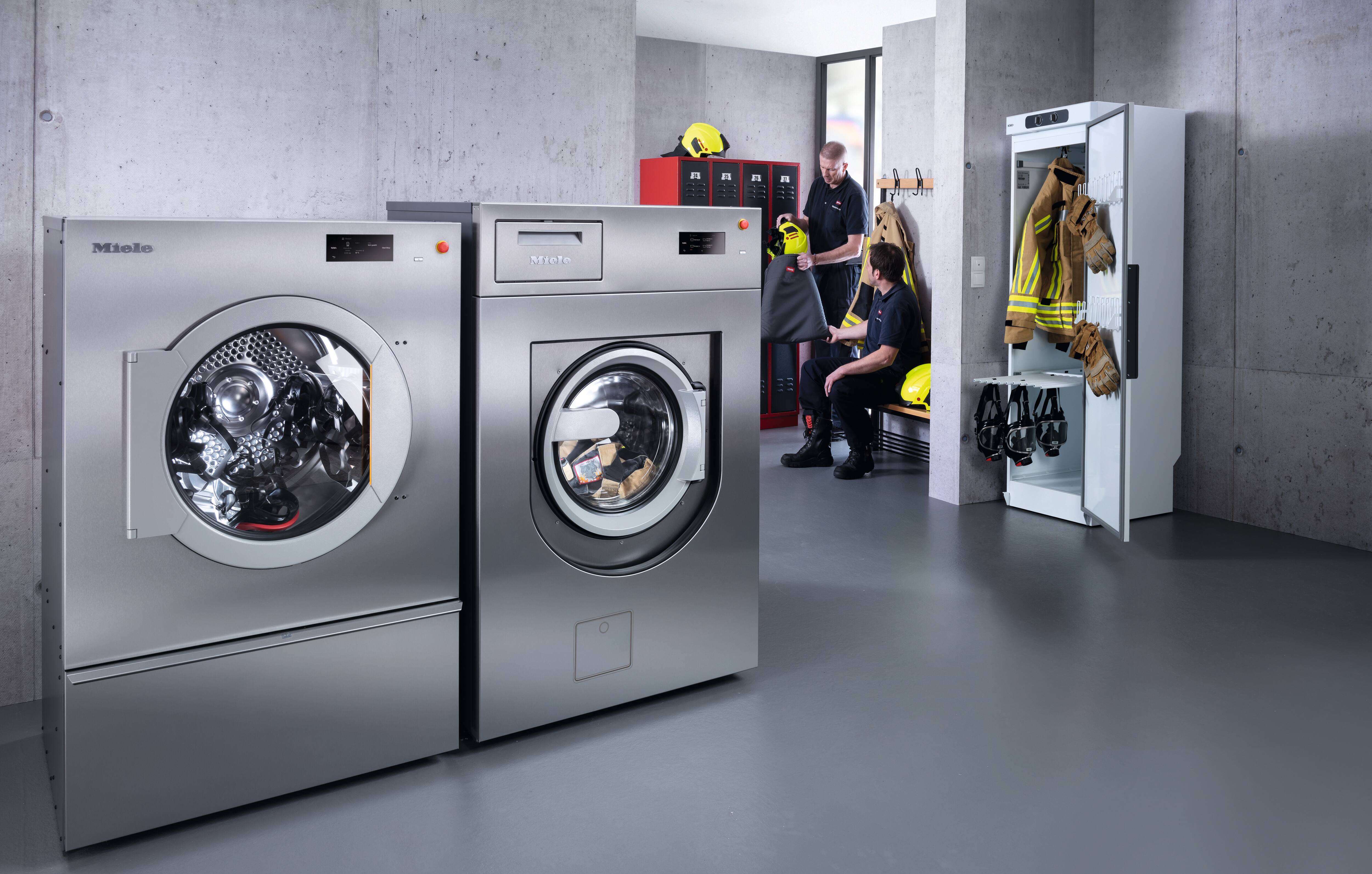 In einer Feuerwehr steht eine gewerbliche Benchnmark Performance Plus PowerWash PRO Waschmaschine rechts neben einem Benchmark Performance Plus Trockner. Ebenfalls im Raum ist ein Trockenschrank zu sehen und im Hintergrund zwei Feuerwehrmänner die sich unterhalten.