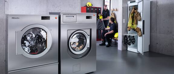 In einer Feuerwehr steht eine gewerbliche Benchnmark Performance Plus PowerWash PRO Waschmaschine rechts neben einem Benchmark Performance Plus Trockner. Ebenfalls im Raum ist ein Trockenschrank zu sehen und im Hintergrund zwei Feuerwehrmänner die sich unterhalten.