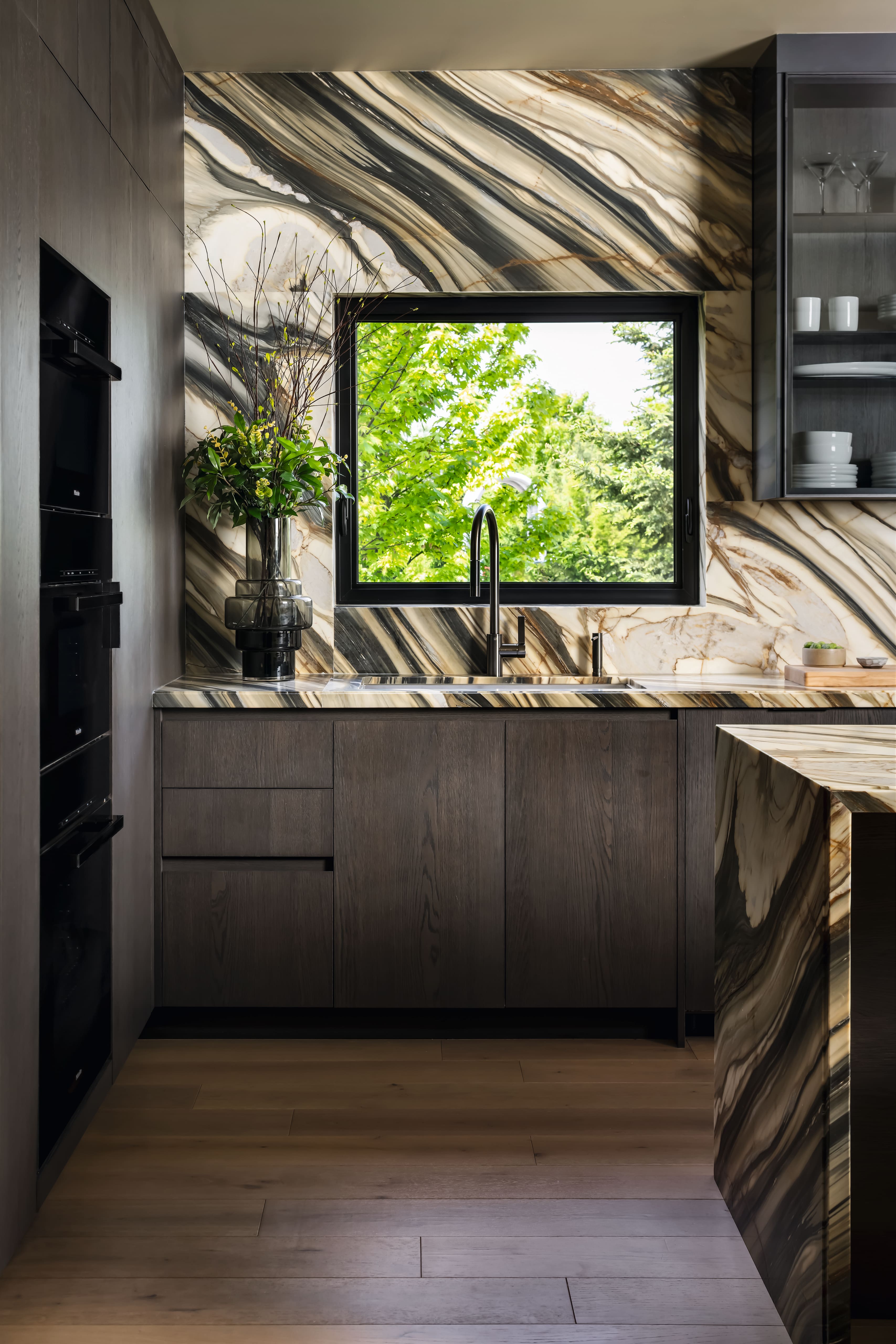 modern kitchen with dark wood cabinetry, a marble-patterned backsplash, and a window framing green trees.