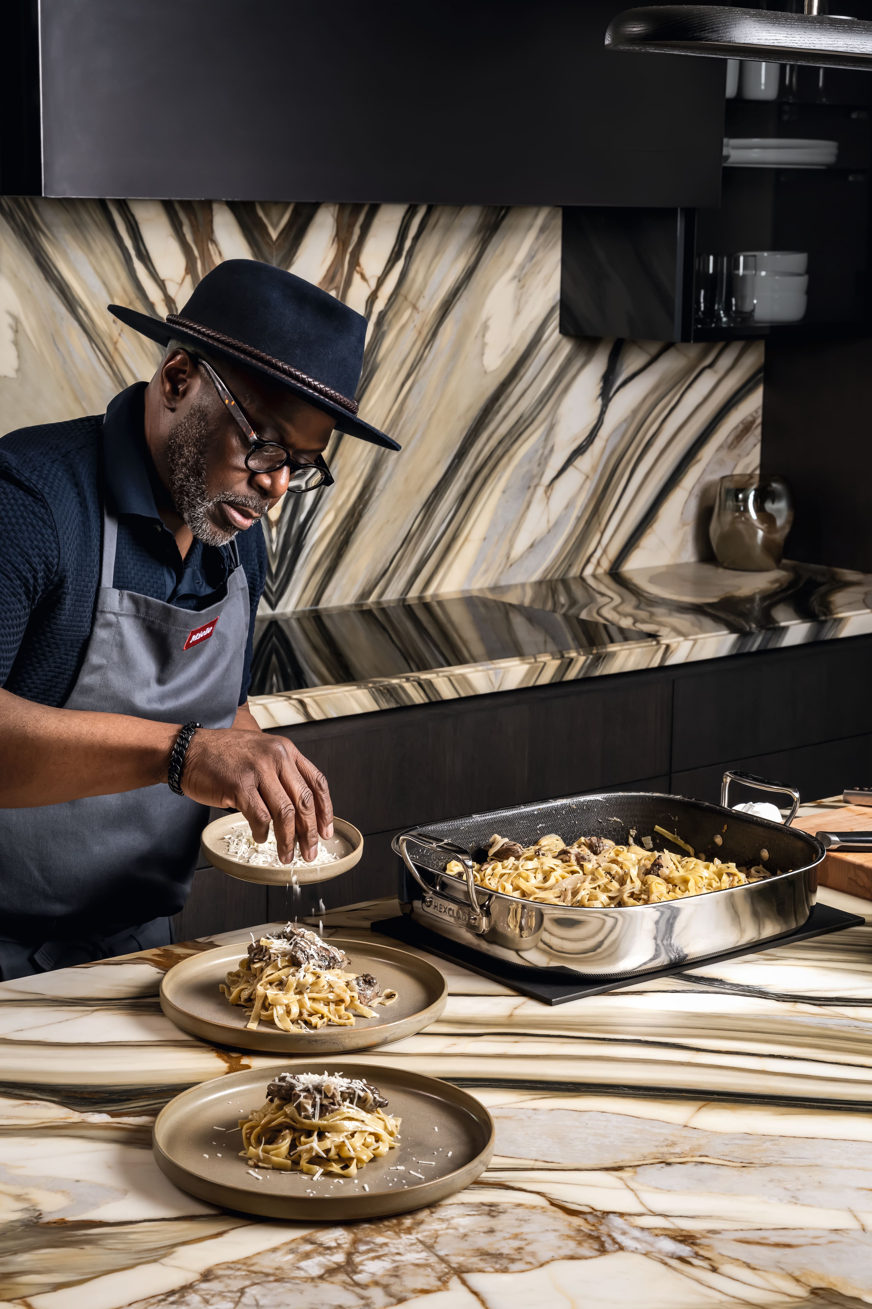 A person wearing a hat and apron stands in a modern kitchen with dramatic marble-patterned surfaces.