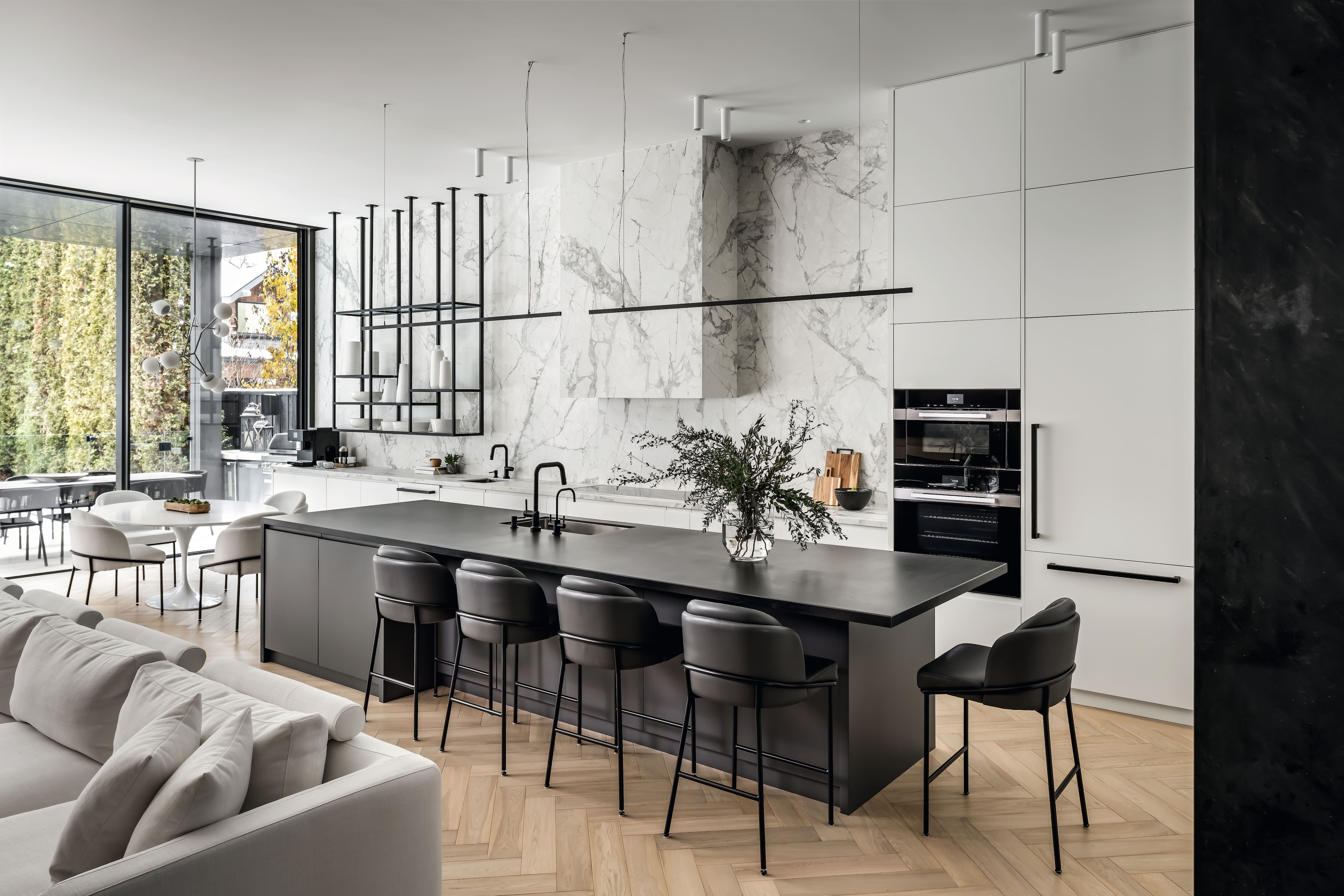 A modern kitchen with a long dark island, black barstools, and marble backsplash extending to the ceiling. The space features sleek white cabinetry, minimalist lighting, and large windows opening to an outdoor dining area