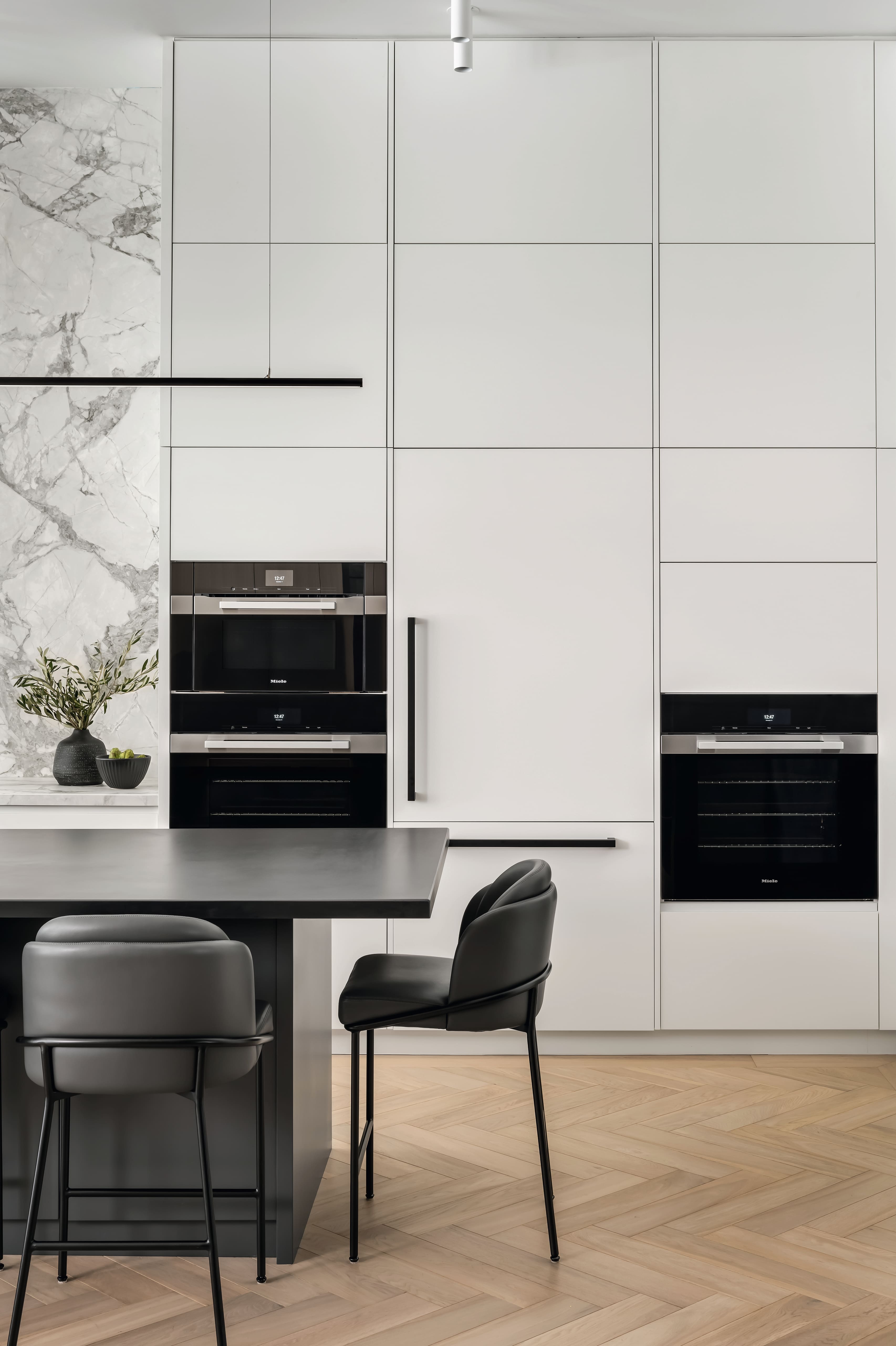 A sleek modern kitchen with white paneled cabinetry, black built-in appliances, and a marble accent wall.