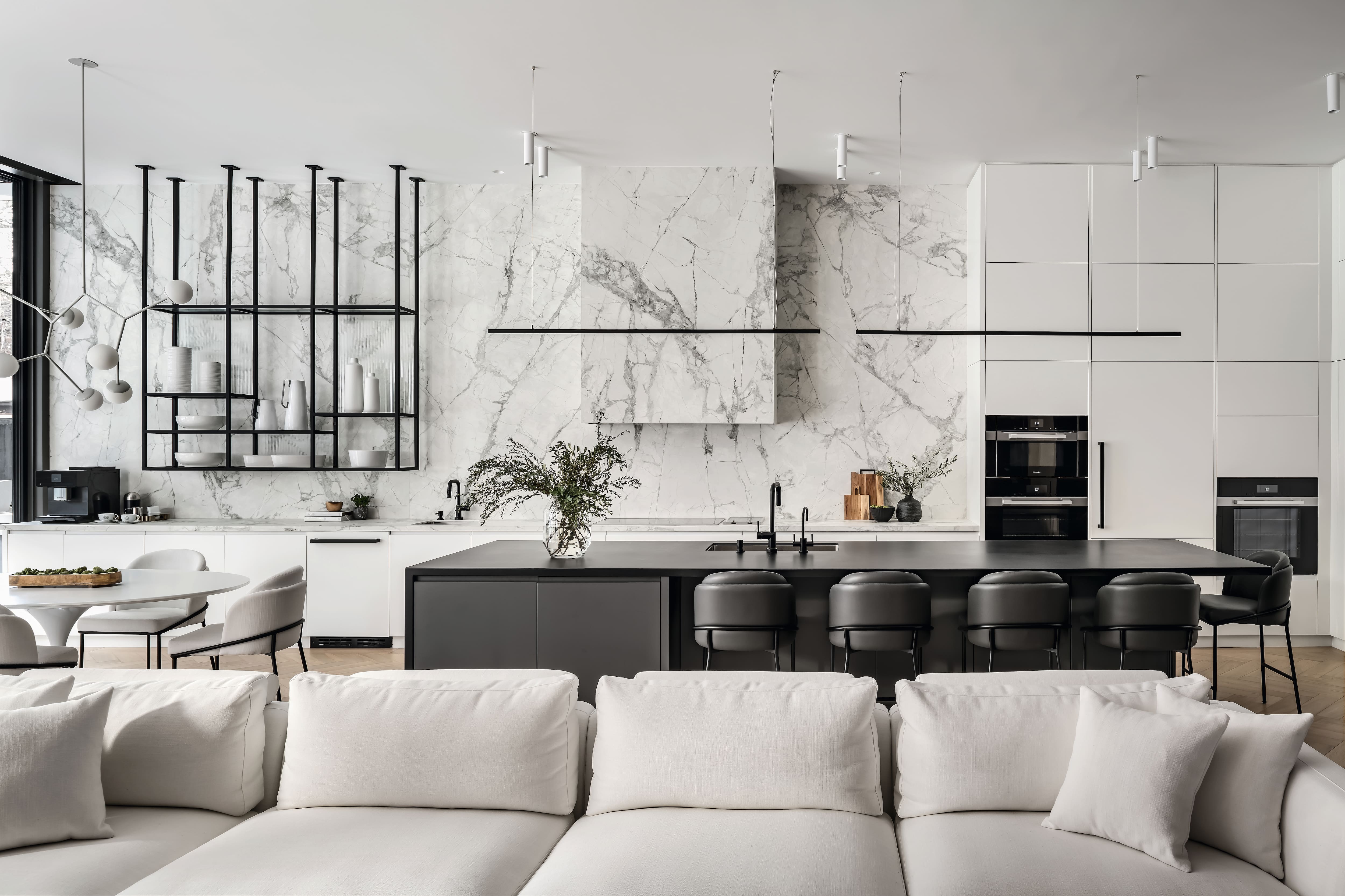 A spacious modern kitchen with a large marble backsplash, dark island seating, and white sofas in the foreground.