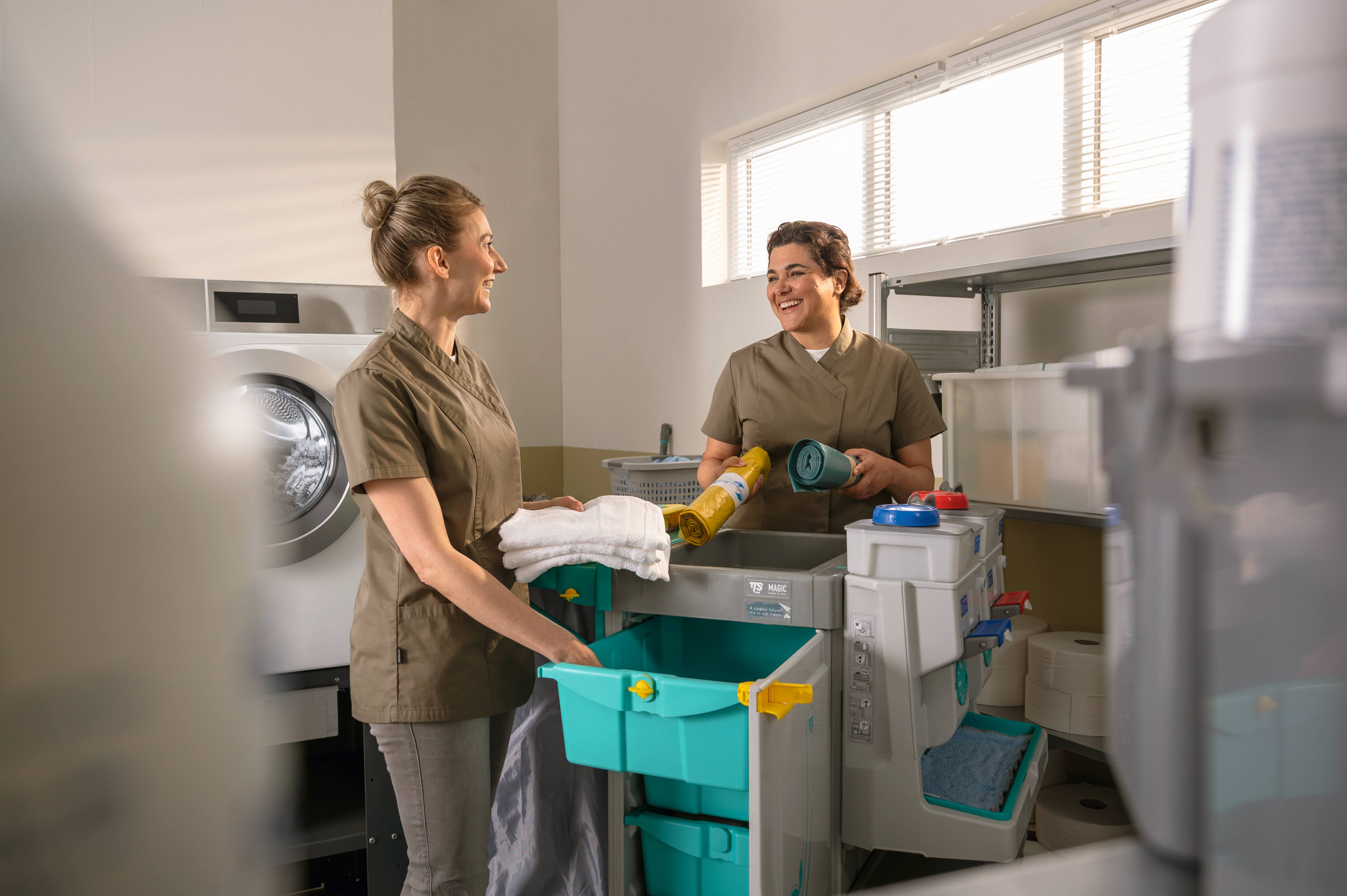 Deux personnes au travail avec des ustensiles de nettoyage et des chiffons à tout usage sur un chariot dans un environnement de laverie professionnelle. 