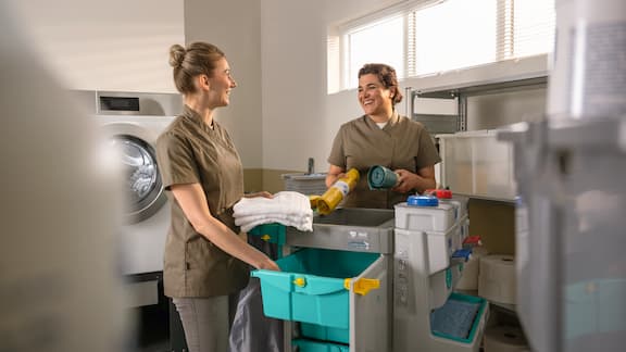 Deux personnes au travail avec des ustensiles de nettoyage et des chiffons à tout usage sur un chariot dans un environnement de laverie professionnelle.