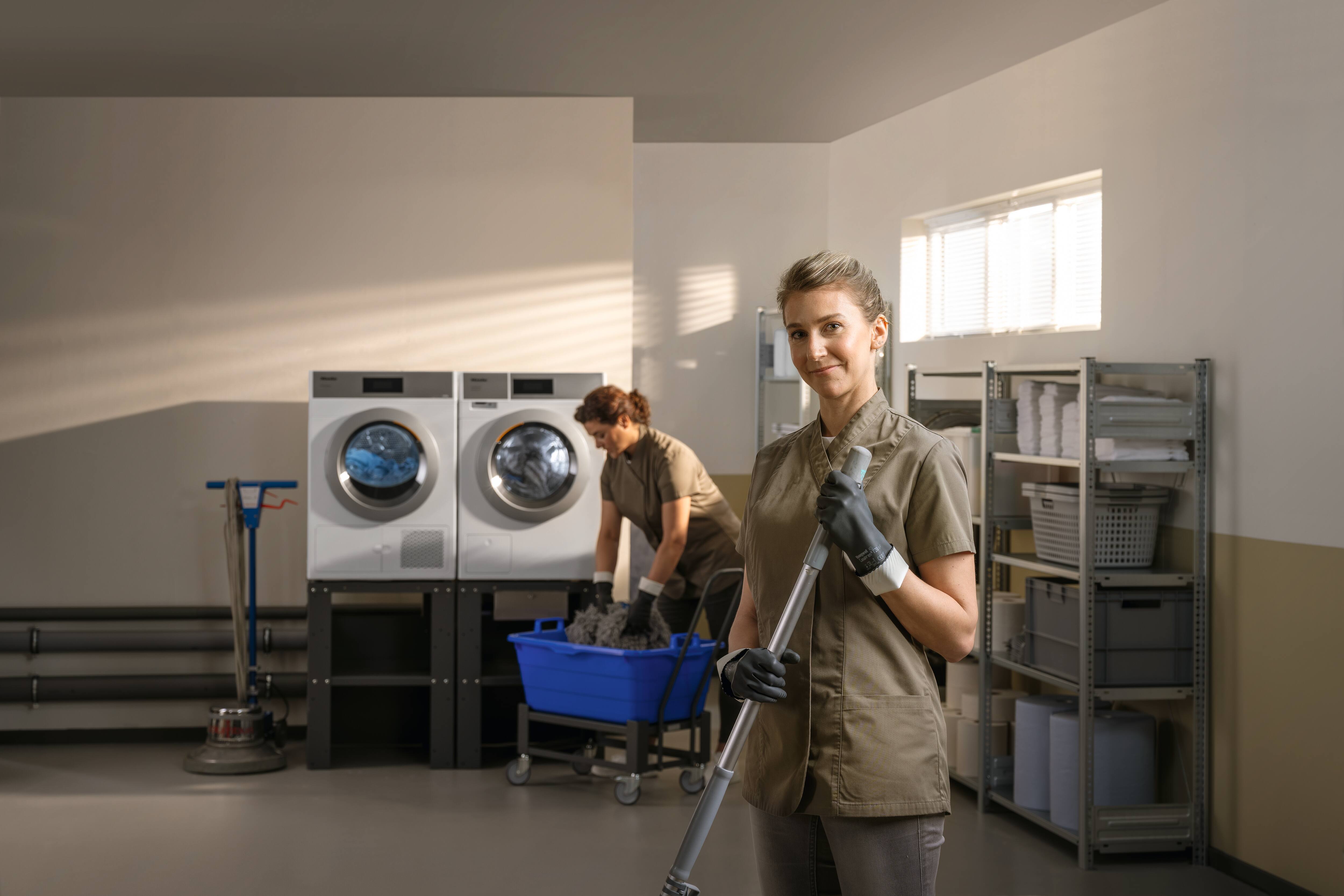 Dans une buanderie, une personne travaillant pour une entreprise de nettoyage se tient debout, un faubert à la main, et sourit à l’objectif. À l’arrière-plan, une autre charge des fauberts dans une laveuse Mop Star de la gamme Petits Géants (Little Giants) de Miele. Une sécheuse Petits Géants (Little Giants) PDR 508 de Miele se trouve à côté de la laveuse.