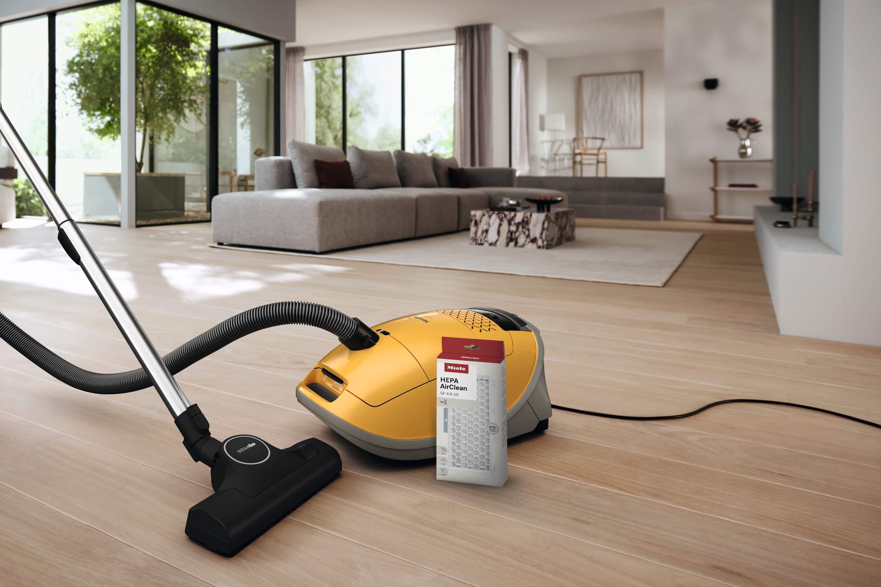 Zdjęcie odkurzacza workowego Miele Guard M1 razem z opakowaniem filtra HEPA do odkurzacza, które stoją na podłodze w nowoczesnym wnętrzu 