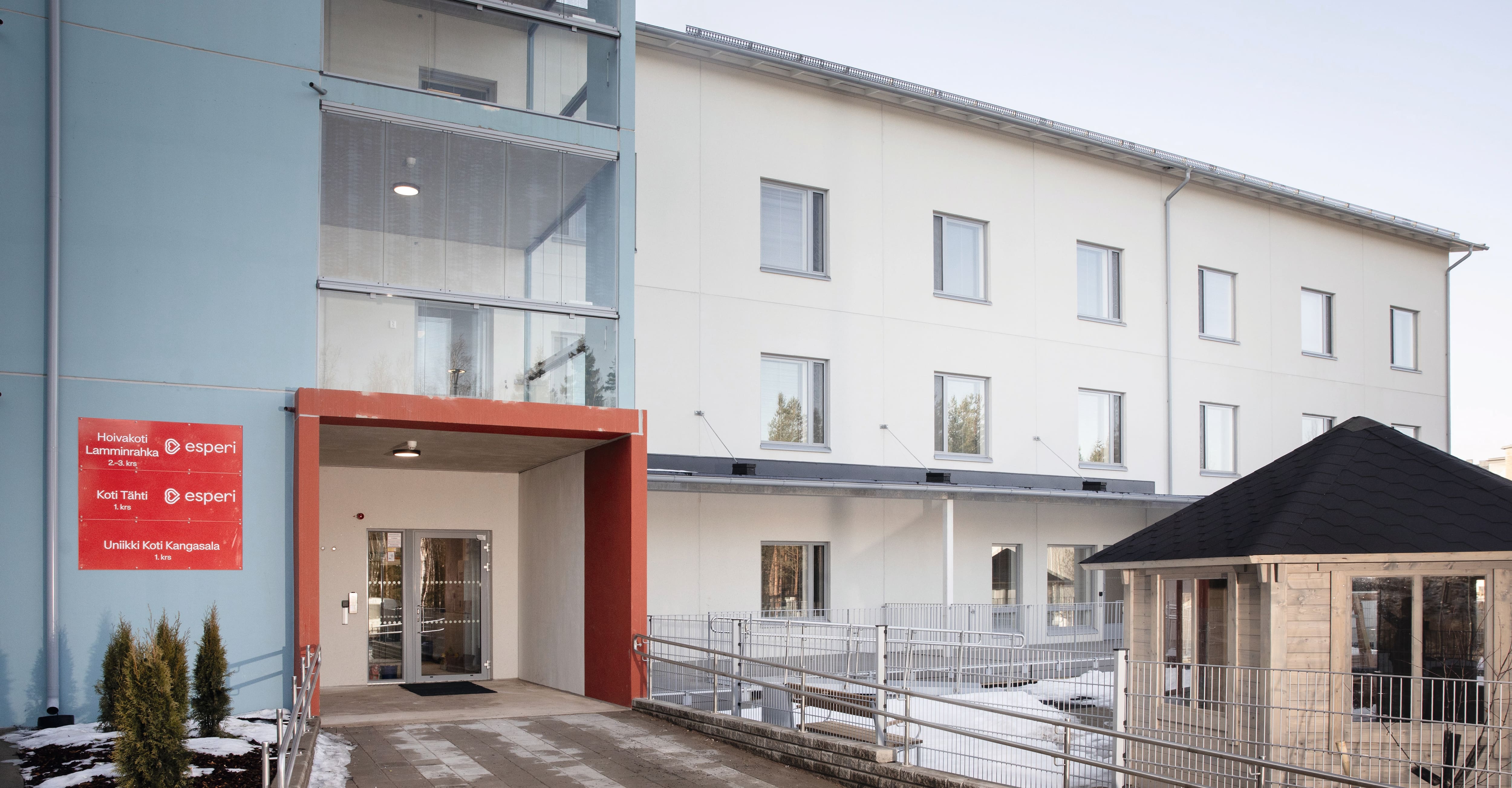 Esperi Hoivakoti Lamminrahkan punaisin reunuksin oleva pääsisäänkäynti ja kolmikerroksiseen vaaleaan taloon. Etualalla ulkoilupuiston kotamökki.